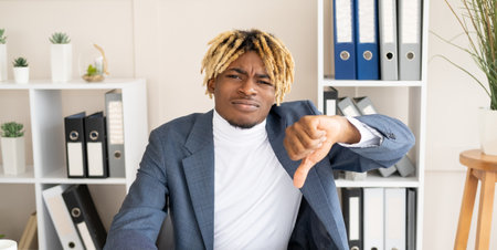 Bad job. Displeased black man. Negative result. Disappointed male worker sitting office desk showing thumb down dislike gesture in light room interior.の写真素材