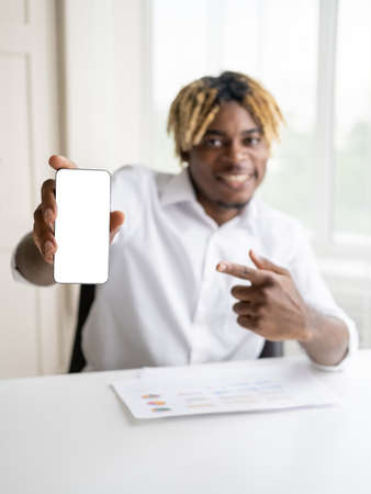 Mobile application. Happy black man. Advertising promotion. Smiling defocused handsome guy pointing smartphone blank screen sitting desk in light room interior.の写真素材