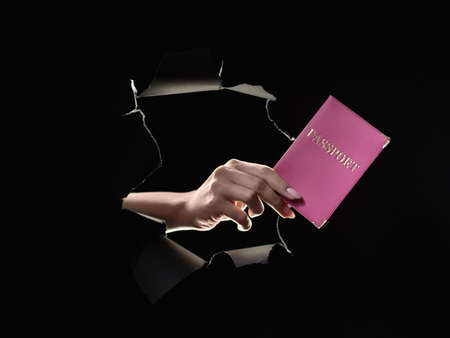 Passport service. Travel document. Global immigration. Female hand holding national ID in pink cover inside black breakthrough hole ripped wall isolated on dark.の写真素材