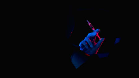 Vaccine shot. Drug injection. Covid-19 jab. Red blue neon light doctor hand in glove holding syringe dose inside black breakthrough hole torn wall isolated on dark copy space banner.の写真素材