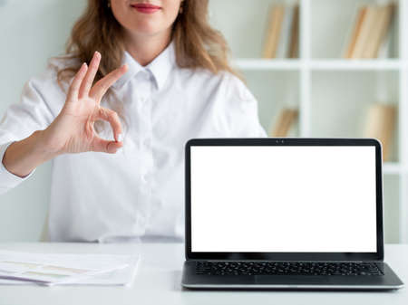 Ok gesture. Official woman. Online education. Pretty unrecognizable smiling lady showing approving sign opened laptop blank screen in light room interior defocused.の写真素材