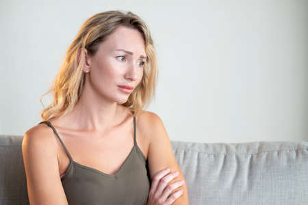 Negative news. Upset woman. Depressed emotion. Sad beautiful lady feeling sorrow almost crying sitting sofa in light room interior.の写真素材