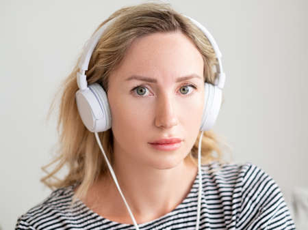 Upset woman. Sound healing. Depressed feelings. Sad worried lady listening in headphones sitting sofa in light room interior.の写真素材