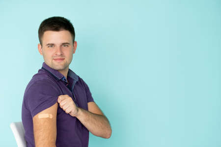 Covid protection. Happy male patient. Vaccination process. Satisfied man showing plaster on shoulder after injection isolated blue copy space.の写真素材