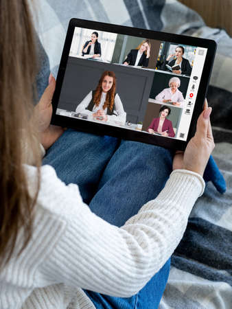 Group video call. Remote meeting. Wfh telecommuting. Relaxed woman using tablet on couch working from home online with female business team on screen.の写真素材