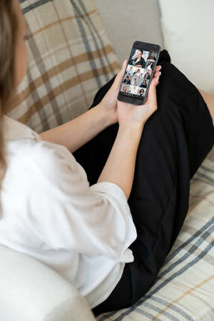 Group video call. Remote work. Mobile telecommuting. Relaxed female freelancer using phone on couch to cooperate with business team online from home.の写真素材