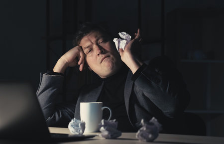 Vertical video. Work crisis. Bored man. Night office. Relaxed lazy male employee messing around throwing creased paper balls in dark room interior.の写真素材