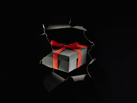 Black Friday. Night sale. Giveaway promotion. Wrapped gift box with red ribbon inside breakthrough torn paper wall isolated on dark empty space commercial background.の写真素材