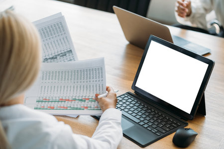 Office work. Female manager. Computer mockup. Unrecognizable woman studying data graphics sitting desk opened laptop blank screen in light room interior.の写真素材