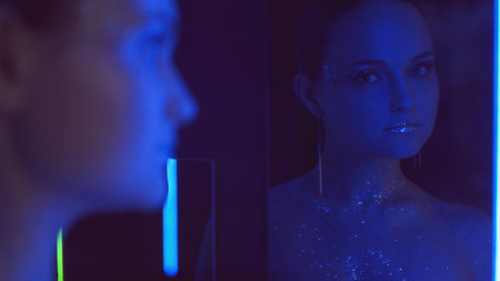 Disco makeup. Neon portrait. Nightclub beauty. Blue purple color light woman with sparkling glitter face skin metallic lips in mirror on dark.の写真素材