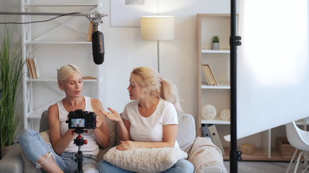 Video blog. Home interview. Blogger communication. Two relaxed women on couch talking recording content on camera on tripod in modern equipped home studio.の写真素材