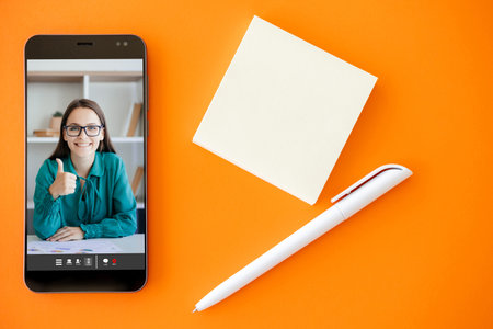 Online expert. Video coaching. Mobile conference. Satisfied business woman approving with thumb up like gesture on phone screen on orange copy space background with pen empty note.の写真素材