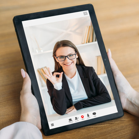 E-learning course. Video chat. Digital education. Female student watching online satisfied teacher approving result with okay on tablet screen at workplace.の写真素材