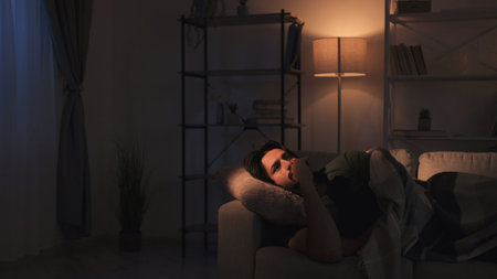 Restless night. Insomnia problem. Mental overload. Sad depressed pensive man overthinking lying on couch late at dark home interior.の写真素材