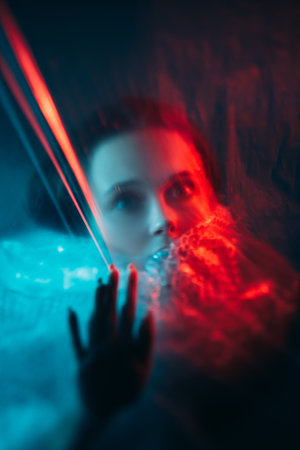 Horror fear. Night terror. Spooky mystery. Red blue color light defocused scared terrified woman wrapped in polyethylene film trapped behind plastic transparent wall on dark.の写真素材