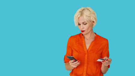 Online payment. Confused woman. Digital technology. Upset puzzled lady holding credit card looking smartphone posing blue copy space.の写真素材