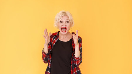 Angry quarrel. Arguing woman. Rage expression. Grimacing middle-aged shouting lady gesturing hands up posing yellow.の写真素材