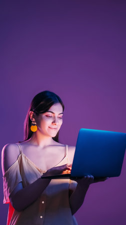 Laptop entertainment. Satisfied calm neon color light girl using computer social network chatting online isolated on purple background.の写真素材