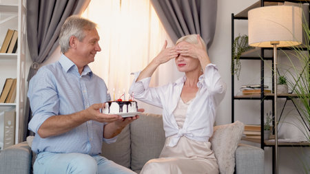Birthday surprise. Happy couple. Holiday celebration. Cheerful middle-aged man presenting festive cake to wife closed eyes sitting light home interior.の写真素材