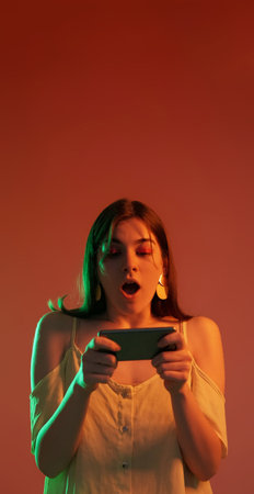 Smartphone playing. Amazed woman portrait. Surprised shocked neon color light girl playing online games isolated on red copy space background.の写真素材