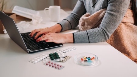 Distance work. Sick woman. Home isolation. Unrecognizable lady wrapped plaid typing laptop on desk with medicine pills in light room interior.の写真素材
