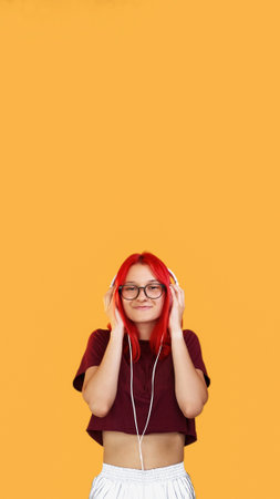 Music headset. Sound technology. Listen pleasure. Young happy redhead teenage girl in eyewear wearing modern white wire headphones on orange background empty space.の写真素材