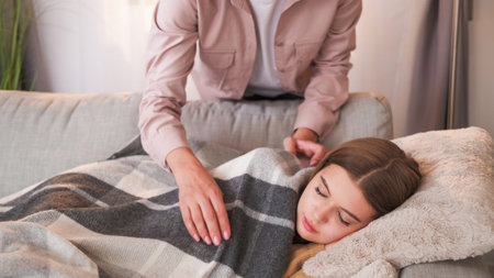 Home nap. Parent care. Unrecognizable woman covering plaid teenager girl sleeping on sofa weekend drowsing in light room interior.の写真素材
