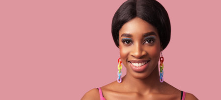 Friendly face. Bright makeup. Summer look. Happy cheerful satisfied smiling woman with colorful rainbow earrings isolated on pink empty space background.の写真素材