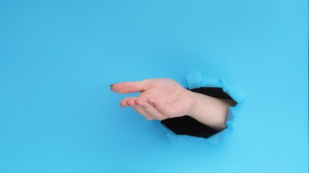 Give gesture. Pay money. Female hand open palm inside breakthrough hole on blue ripped paper wall advertising background with empty space.の写真素材