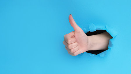 Like gesture. Positive feedback. Female hand showing approval thumb up on blue torn breakthrough paper hole background with empty space.の写真素材