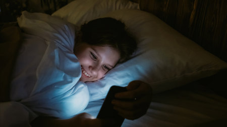Kid addiction. Digital leisure. Night internet surfing. Happy smiling girl scrolling social media feed on smartphone lying in bed in dark room at home.の写真素材