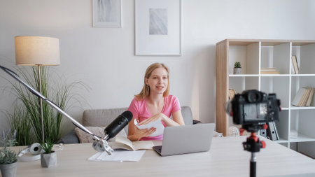 Coaching blog. Content recording. Confident smiling young woman at table with notes talking to camera on tripod and microphone filming video in light room.の写真素材
