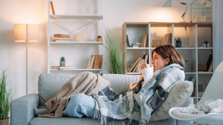 Sick tea. Virus drinking. Sore throat. Shivering woman catching cold suffering influenza holding cup healing grippe sitting wrapped blanket on couch in living room at home.の写真素材