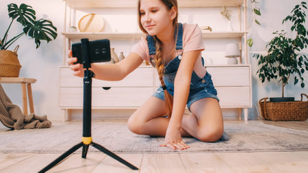 Kid vlog. Blogger home lifestyle. Create content. Happy smiling child girl adjusting phone camera on tripod recording video in light interior.の写真素材