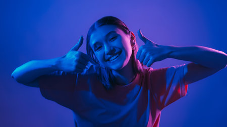 Like gesture. Cool offer. Positive recommendation. Happy smiling woman in vibrant neon light showing thumbs up on blue background.の写真素材