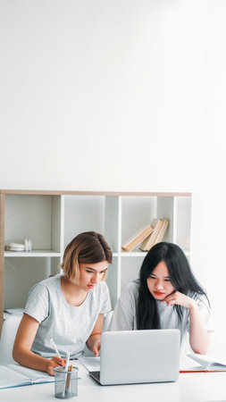 Studying together. Homework cooperation. Two young girls at desk with laptop learning completing task preparing for exam writing notes at home empty space.の写真素材