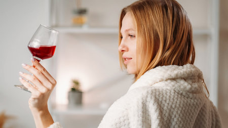 Spa alcohol. Weekend drink. Vacation toast. Blonde woman in bathrobe with glass of red wine on light...