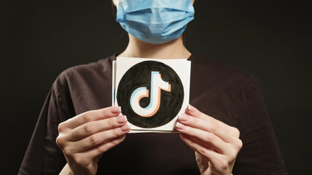 Kharkiv, Ukraine - May 12, 2020: Social network quarantine. Pandemic content. Unrecognizable influencer woman in face mask showing TikTok logo on dark black copy space.のeditorial素材
