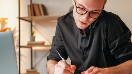 Remote management. Mobile communication. Smart business man in glasses working from home speaking on phone taking notes at modern office workplace interior with free space.の写真素材