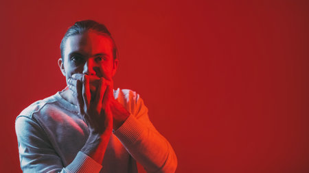 Keep quiet. Silence warning. Conspiracy secret. Red color neon light disturbed man hands covering mouth on dark empty space background.の写真素材