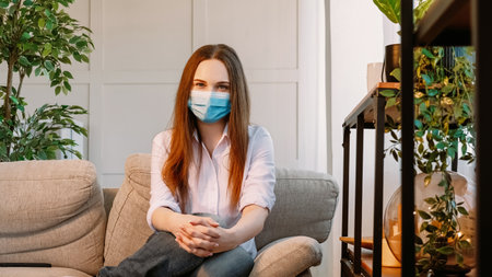 Home isolation. Quarantine hygiene. Beautiful woman in protective medical mask spending time alone during pandemic siting on sofa in cozy light home interior.の写真素材
