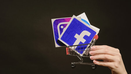 Kharkiv, Ukraine - May 12, 2020: Social media marketing. Network promotion. Female hand holding Facebook Instagram icon in shopping cart on dark black empty space background.のeditorial素材