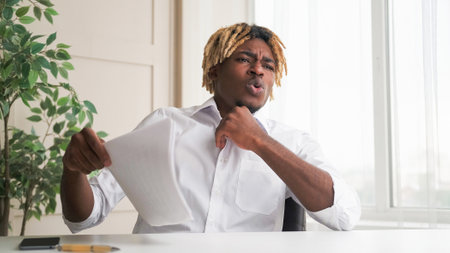 Hot office. Broken heat system. Exhausted business man suffering from boiling day sitting at desk waving blowing wind with documents at modern interior workplace.の写真素材