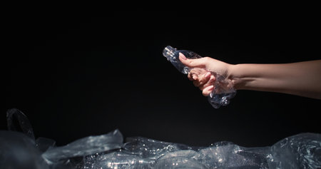 Waste reduction. Plastic recycling. Woman volunteer hand crushing used bottle throwing into trash dump with cellophane bags isolated on black background empty space.の写真素材