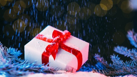 Christmas celebration. Holiday gift composition. Surprise present box wrapped white paper with red ribbon bow on pine branch xmas decor snow falling magic lights background.の写真素材