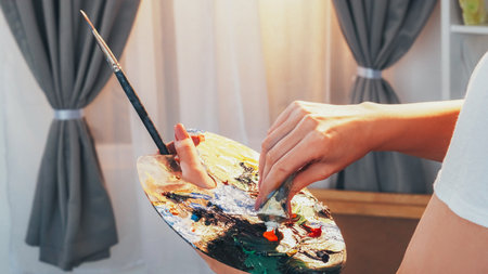 Art process. Creating artwork. Closeup of woman painter hand mixing oil colorful paint on messy palette with paintbrush at artist studio.の写真素材
