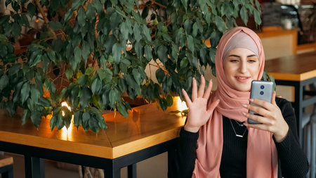 Online stream. Video communication. Cheerful woman in hijab waving hand to followers greeting subscribers social media content at cozy cafe.の写真素材