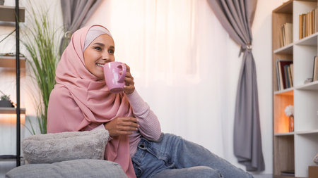 Home coffee. Weekend leisure. Happy woman in hijab with mug enjoying hot drink at light modern...