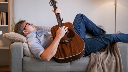 Musician relax. Home rest. Peaceful casual dreamy man laying on sofa playing acoustic guitar composing melody in light cozy living room interior.の写真素材