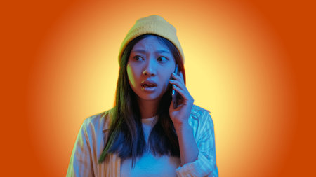 Phone scam. Mobile fraud. Blue color neon light overwhelmed hipster girl talking using gadget isolated on orange gradient copy space background.の写真素材
